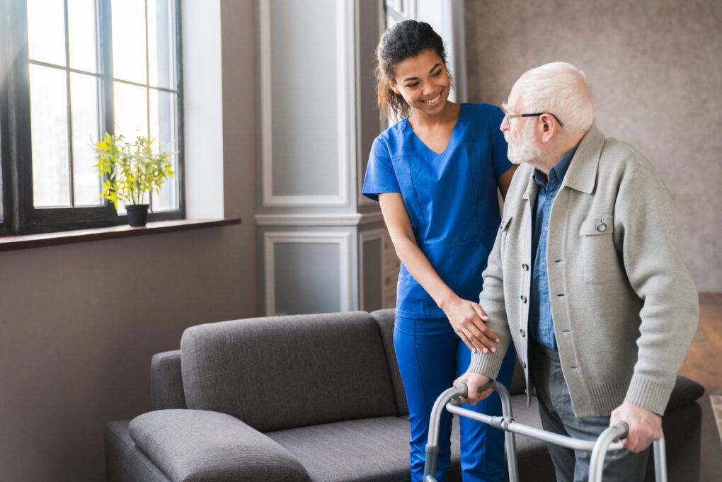 Portrait of a young nurse helping elderly man grandfather to walk using walker equipment in the bedroom. Senior patient of nursing home moving with walking frame and nurse support